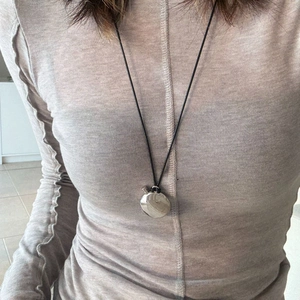 Detail of a long black cord necklace with a polished silver oval pendant on an apricot rib knit