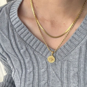Dettaglio di una collana a strati dorata con ciondolo a bussola su maglione grigio