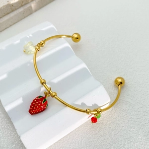 Golden bow cuff bracelet with strawberry and cherry charms on a white textured stand
