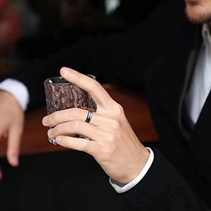Detail eines matt schwarzen Titanrings mit blauem Streifen an einer Hand mit einem Glas