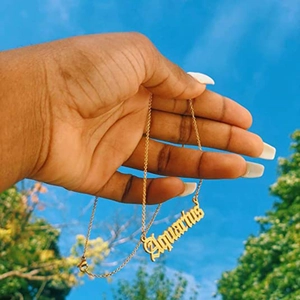 Détail du pendentif Verseau doré en lettres gothiques tenu devant un ciel bleu clair