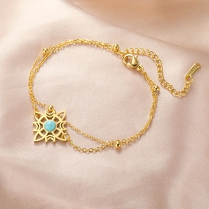 Detail eines goldenen Doppelkettenarmbands mit Sonnenanhänger und Türkis auf rosa Satin