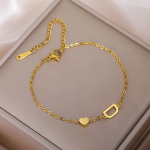 Goldenes Edelstahl-Armband mit Herz- und D-Anhänger auf grauer Schmuckschatulle