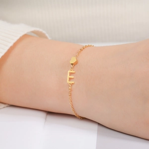 Detail eines goldenen Edelstahl-Armbands mit E-Buchstabe und Herzanhänger am Handgelenk