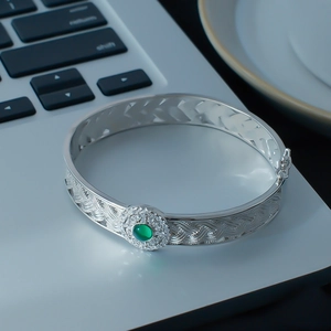 Silver open bangle with hollow lace texture and green stone centerpiece on grey background