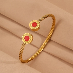 Détail du bracelet en câble doré avec des breloques rouges et un motif grec de méandres
