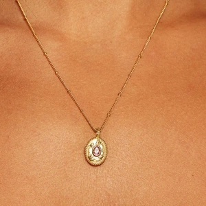 Close-up of a gold locket necklace with floral relief and a pink gemstone on a model