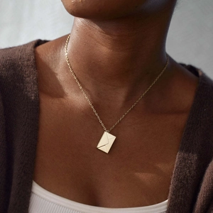 Gouden envelop hanger ketting van titanium staal gedragen door een model op een bruine trui