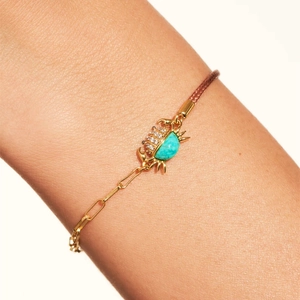 Detalle de una pulsera de cangrejo dorada con turquesa y cuerda tejida en la muñeca