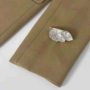 Détail d'une broche feuille argentée plissée sur le revers d'un blazer de couleur camel