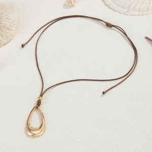 Detalle de colgante de gota dorada en cordón marrón ajustable con fondo de temática playera