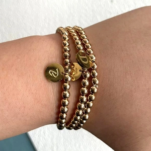 Detailweergave van drie gouden kralenarmbanden met gegraveerde letterhangers om de pols