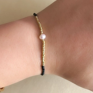 Primer plano de una pulsera minimalista de cuentas doradas, negras y una perla rosa central