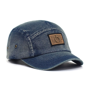 Close-up of washed denim fabric with brown decorative patch and metal ring on the 5-panel cap