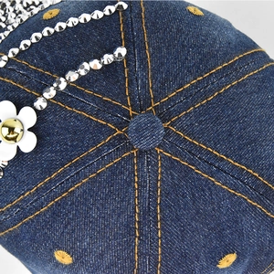 Detail of denim baseball cap showing silver rivets, white flower and yellow contrast stitching