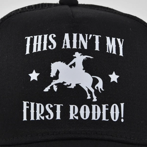 Detail of black trucker hat with white This Ain't My First Rodeo printed cowboy graphic