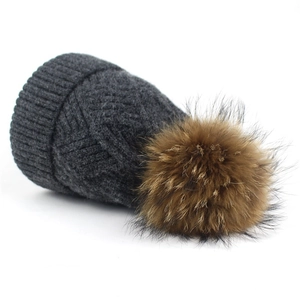 Detail of dark grey knit winter beanie featuring a soft fluffy raccoon fur pompom top