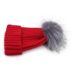 Detailed view of red ribbed knit beanie with a fluffy grey faux fox fur pompom decoration