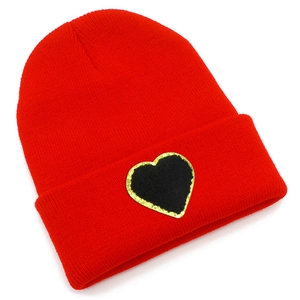 Close-up of red knit beanie featuring a black heart patch with gold sequin border