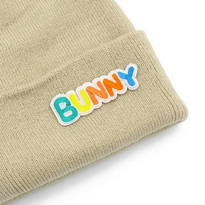 Detail view of beige chunky knit beanie featuring colorful BUNNY letter embroidery patch
