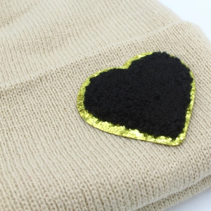Beige knit beanie featuring a heart patch with black plush center and gold sequin trim