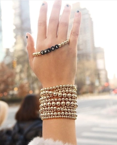Close-up of layered gold beaded bracelets with a black acrylic Chloe name charm bead