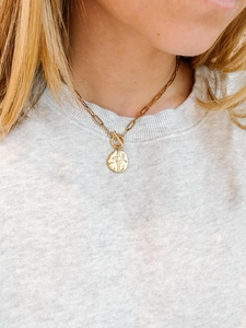 Model wearing a gold paperclip chain necklace with cross coin pendant and toggle clasp