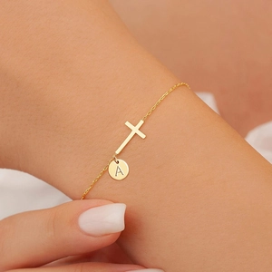 Detail of a gold cross bracelet with a personalized initial A round charm on a delicate chain