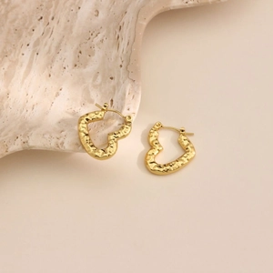Detailed close-up of gold heart hoop earrings with unique hammered texture on beige background