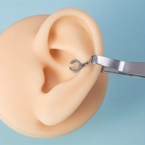 Macro shot showing a stainless steel C-ring being held by tweezers near a silicone ear model