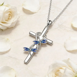 Detalle de collar cruz de plata con libélulas azules sobre mármol y pétalos de rosa blanca