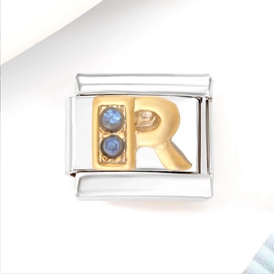 Macro view of stainless steel charm link with gold letter R and blue cubic zirconia stones