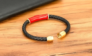 Detalle de pulsera de cuero trenzado negro con cuenta de esmalte rojo y anillos dorados
