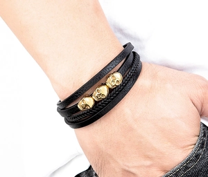 Detail eines mehrlagigen schwarzen Lederarmbands mit drei goldenen Punk-Totenkopf-Anhängern