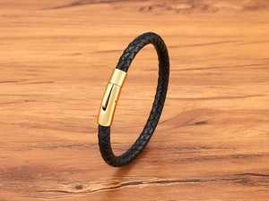 Detail of woven black leather bracelet with a polished gold-tone metal clasp on wood