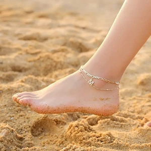 Nahaufnahme der goldenen zweilagigen Fußkette mit Zirkon-Buchstabe M am Sandstrand