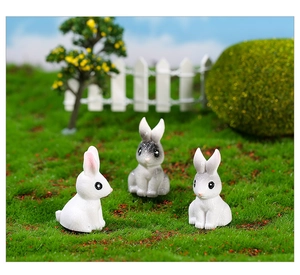 Three miniature resin rabbit figurines on green moss lawn with white picket fence