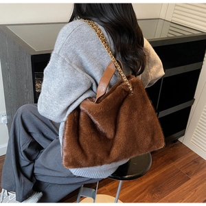 Plush brown faux fur tote bag with gold chain strap shown on a model in a modern interior