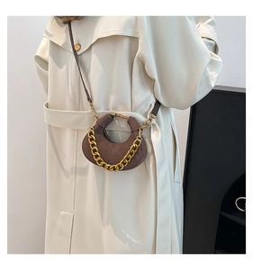 Detalle de un bolso de media luna en ante marrón con cadena dorada y gabardina blanca