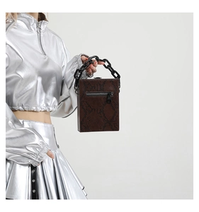 Detail view of brown snakeskin box bag with chunky black chain handle held by a fashion model