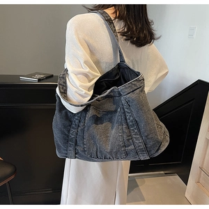 Model wearing a large charcoal denim tote bag with wide strap and stitched pocket detail