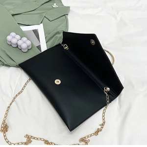 Détail d'une pochette enveloppe noire avec bandoulière chaîne dorée sur fond de tissu blanc