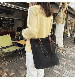 Detail of black canvas tote bag with brown leather handles worn by person in outdoor setting