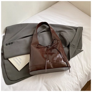 Detail of a dark brown glossy leather tote bag featuring a decorative bow on grey blazer