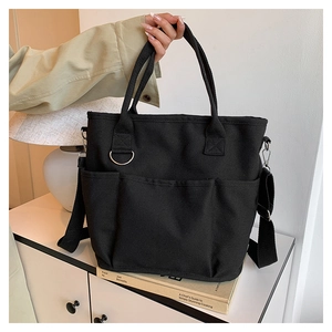 Detail of black canvas tote bag with front pockets and adjustable strap on a white cabinet