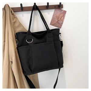 Detailed view of black canvas tote bag with front pockets and D-ring on a wooden hook
