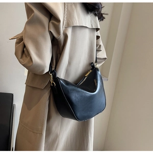 Modern black half-moon crossbody bag in glossy leather worn with a khaki trench coat