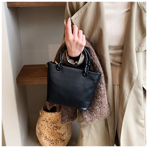 Detail of black pleated bucket bag with round woven handle held by a model in a beige coat