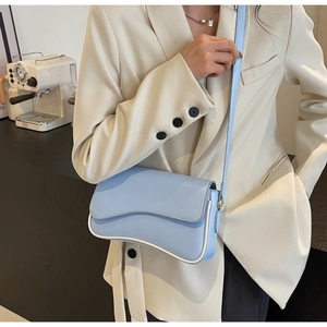 Detail van lichtblauwe crossbodytas met golvende flap en contrastbies op beige blazer