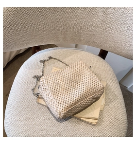 Close-up of a beige straw woven bag with cross-texture and a silver chain on a soft chair